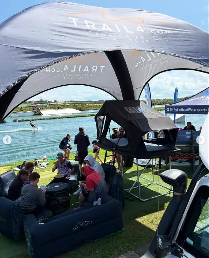 A scenic view from under a "TRAIL4.COM" inflatable tent at a lakeside event, showing a person water skiing in the background, with people eating and chatting on inflatable furniture in the foreground.